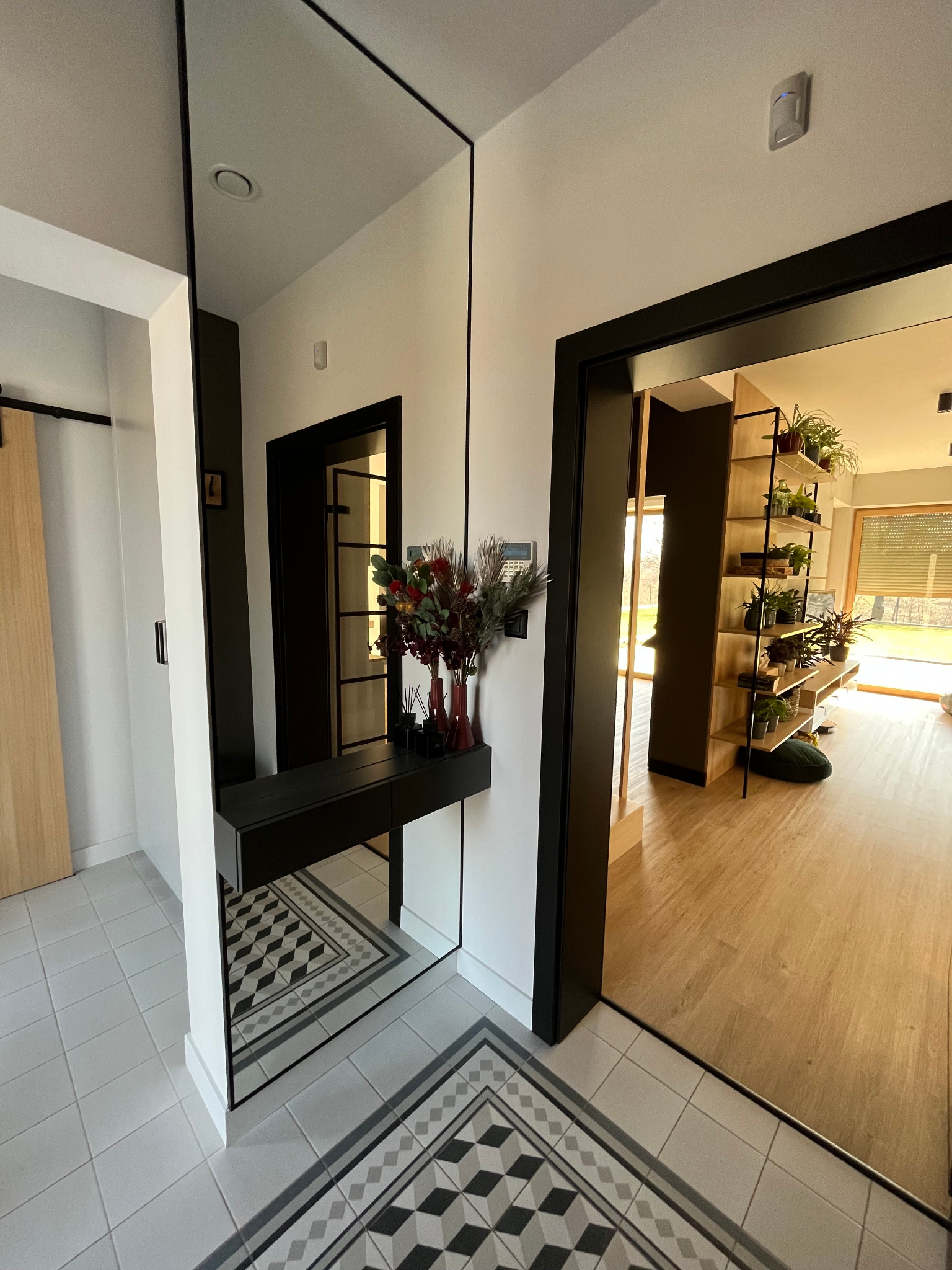 Nowoczesny przedpokój z dużym lustrem i czarną konsolą. Widoczne drzwi z czarną ramą i półki z roślinami w tle. Jasna podłoga z wzorem.