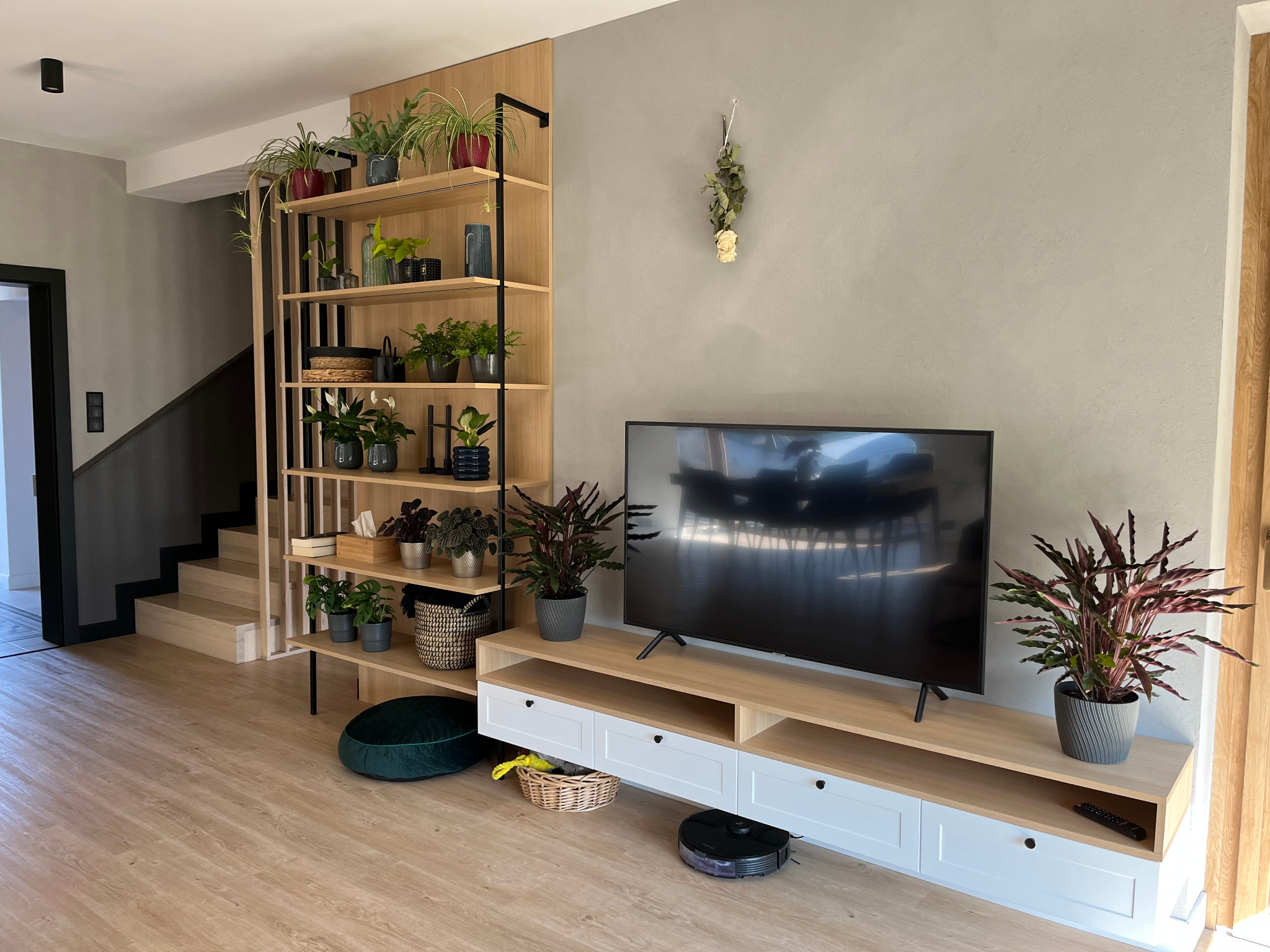 Nowoczesny salon z drewnianą zabudową TV i regałem na rośliny, robot sprzątający. Jasne drewno, szare ściany i podłoga tworzą spójną całość. Minimalistyczny design.