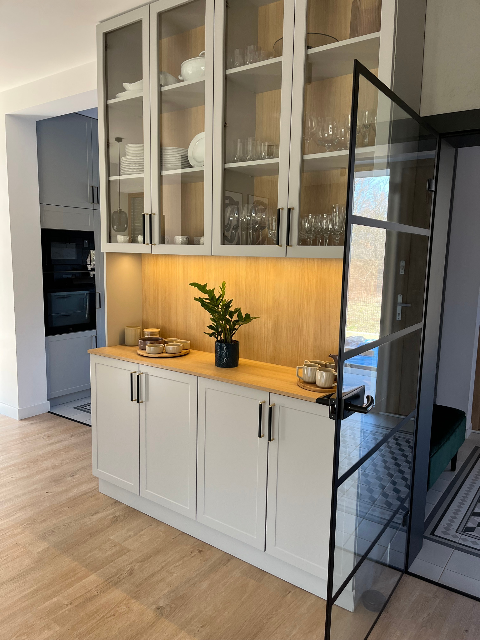 Jasna zabudowa kuchenna z przeszklonymi szafkami i drewnianym blatem, oświetlona od spodu. Na blacie naczynia i roślina w doniczce. Widoczna otwarta, szklana witryna.