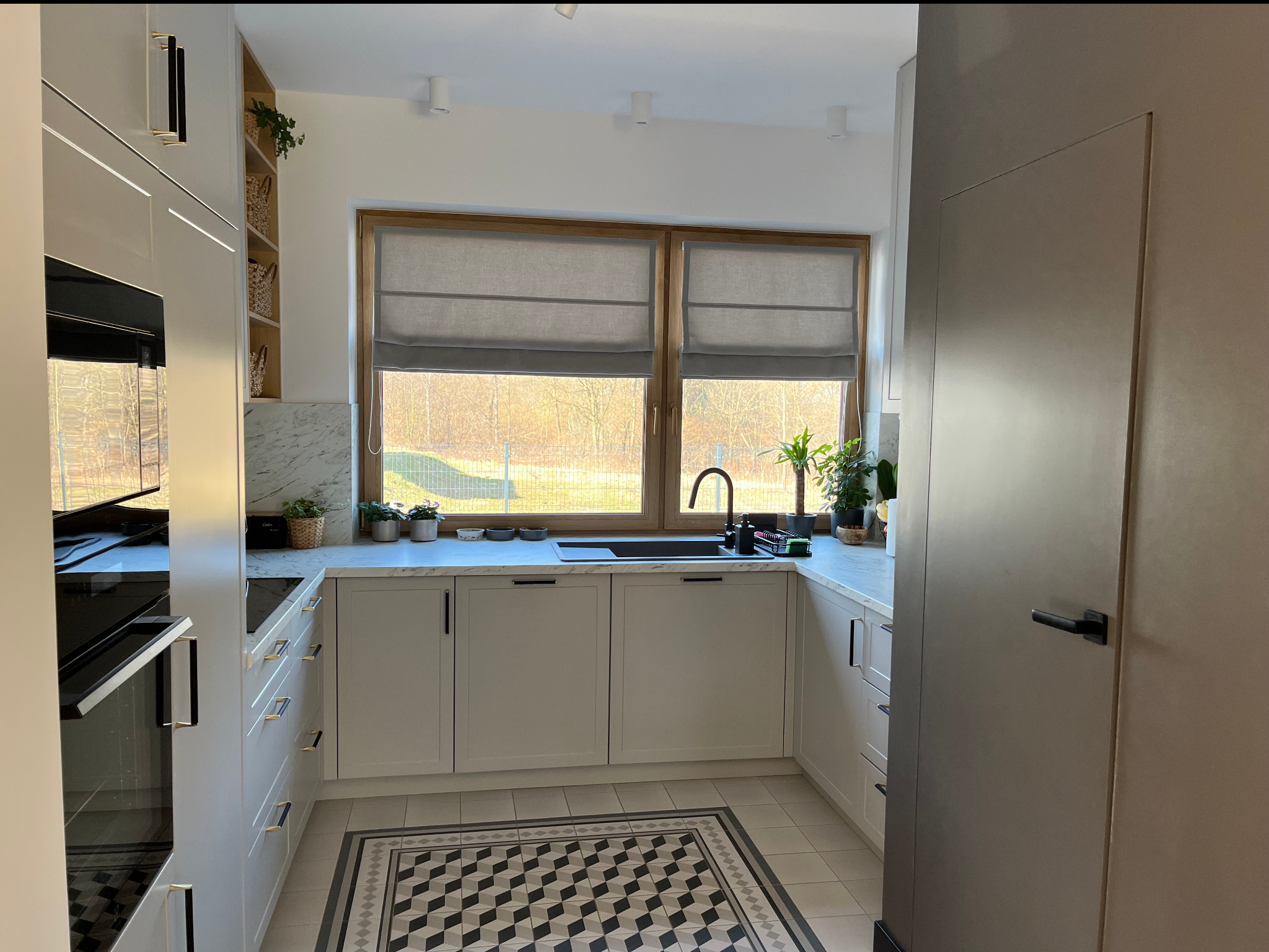 Jasna kuchnia z marmurowym blatem, drewnianą ramą okna i geometryczną podłogą. Rośliny w doniczkach i koszykach dodają naturalnego akcentu.