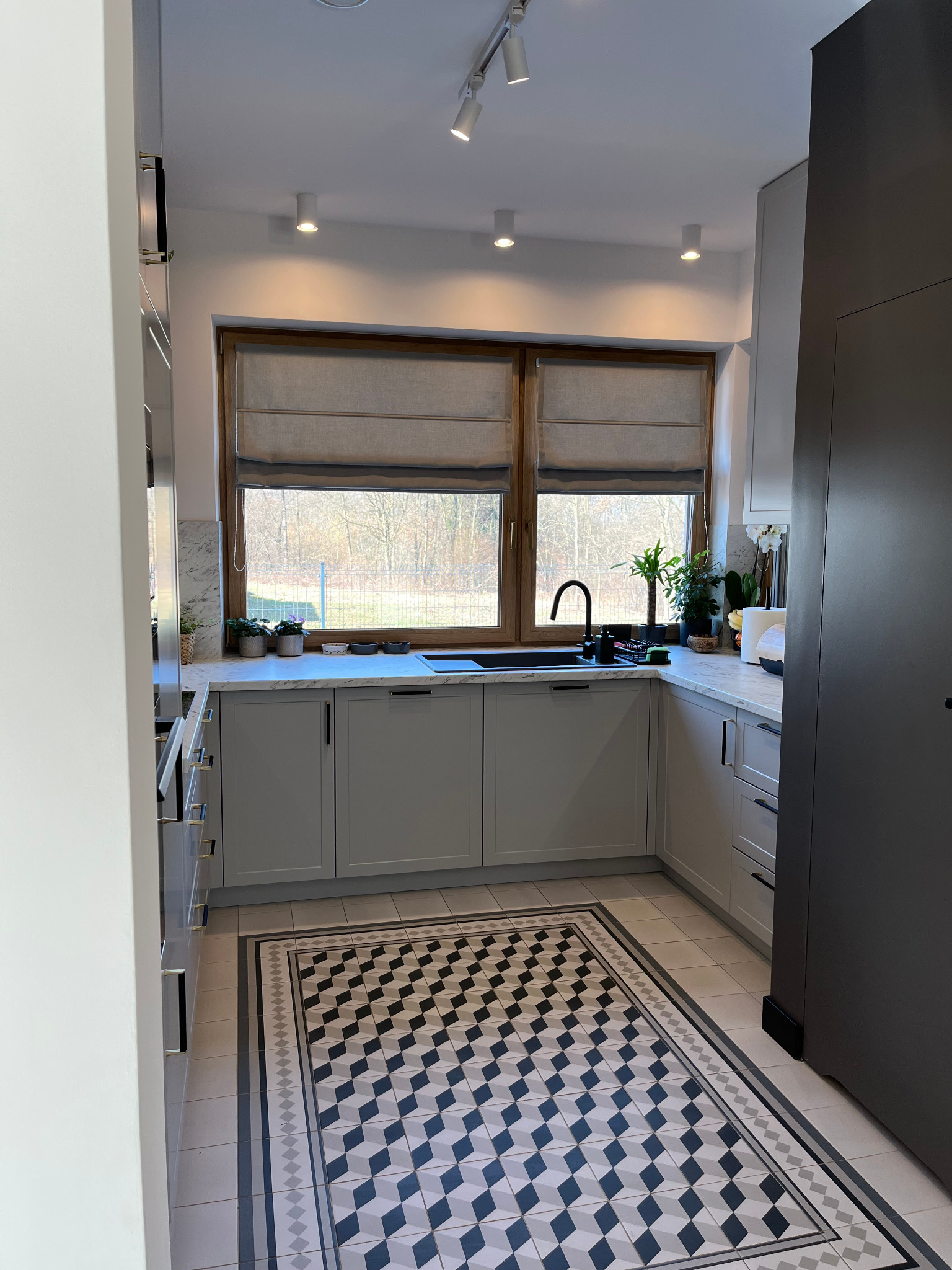 Jasna kuchnia z szarymi szafkami, drewnianym oknem i wzorzystą podłogą w romby. Na blacie marmurkowe wykończenie i rośliny. Nowoczesne oświetlenie sufitowe.