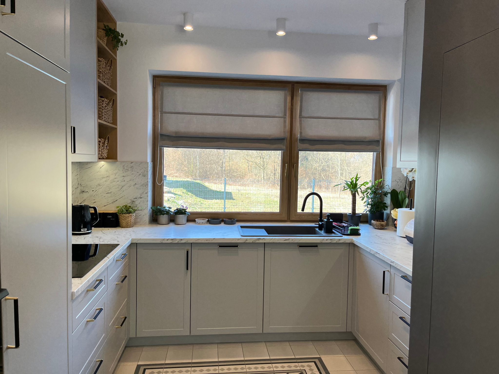 Jasna kuchnia z marmurowym blatem, czarną armaturą i oknem z widokiem na zieleń. Uwagę zwracają minimalistyczne uchwyty szafek i dekoracyjne rośliny.