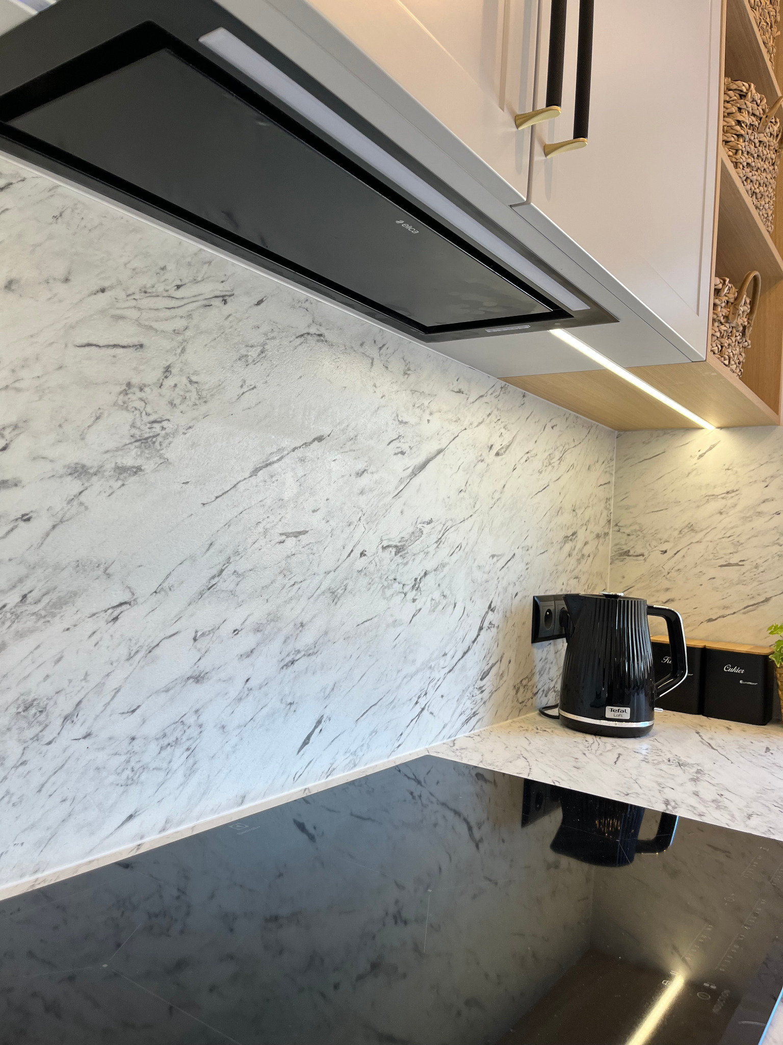 Fragment kuchni z marmurowym blatem, płytą indukcyjną i czajnikiem. Nad blatem szafki z pochłaniaczem i oświetleniem LED. W tle półki z koszykami.
