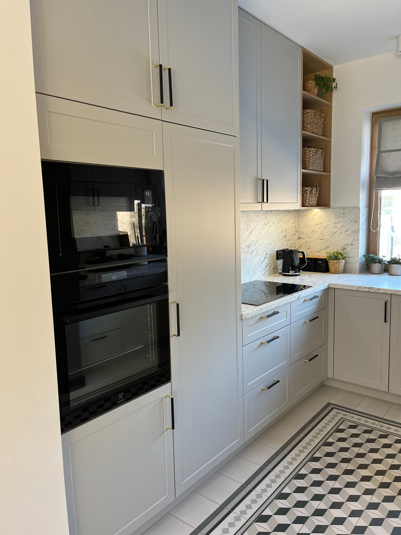 Jasnoszara kuchnia z zabudową po sufit, czarnymi uchwytami i marmurowym blatem. W tle półki z wiklinowymi koszami i roślinami. Nowoczesny design.