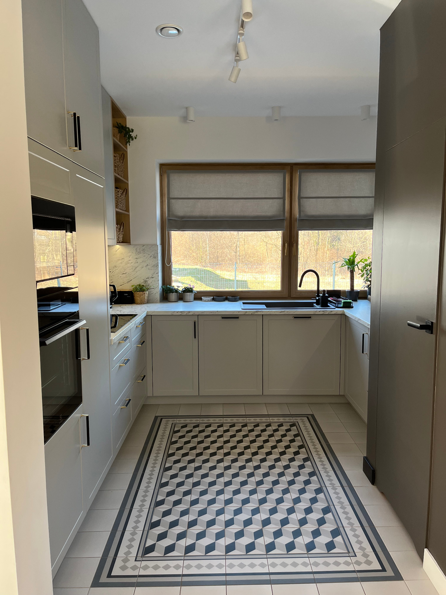 Jasna kuchnia z szarymi frontami, czarną armaturą i geometrycznym dywanem. Widok na okno z roletami rzymskimi i roślinami na parapecie. Minimalistyczny design.