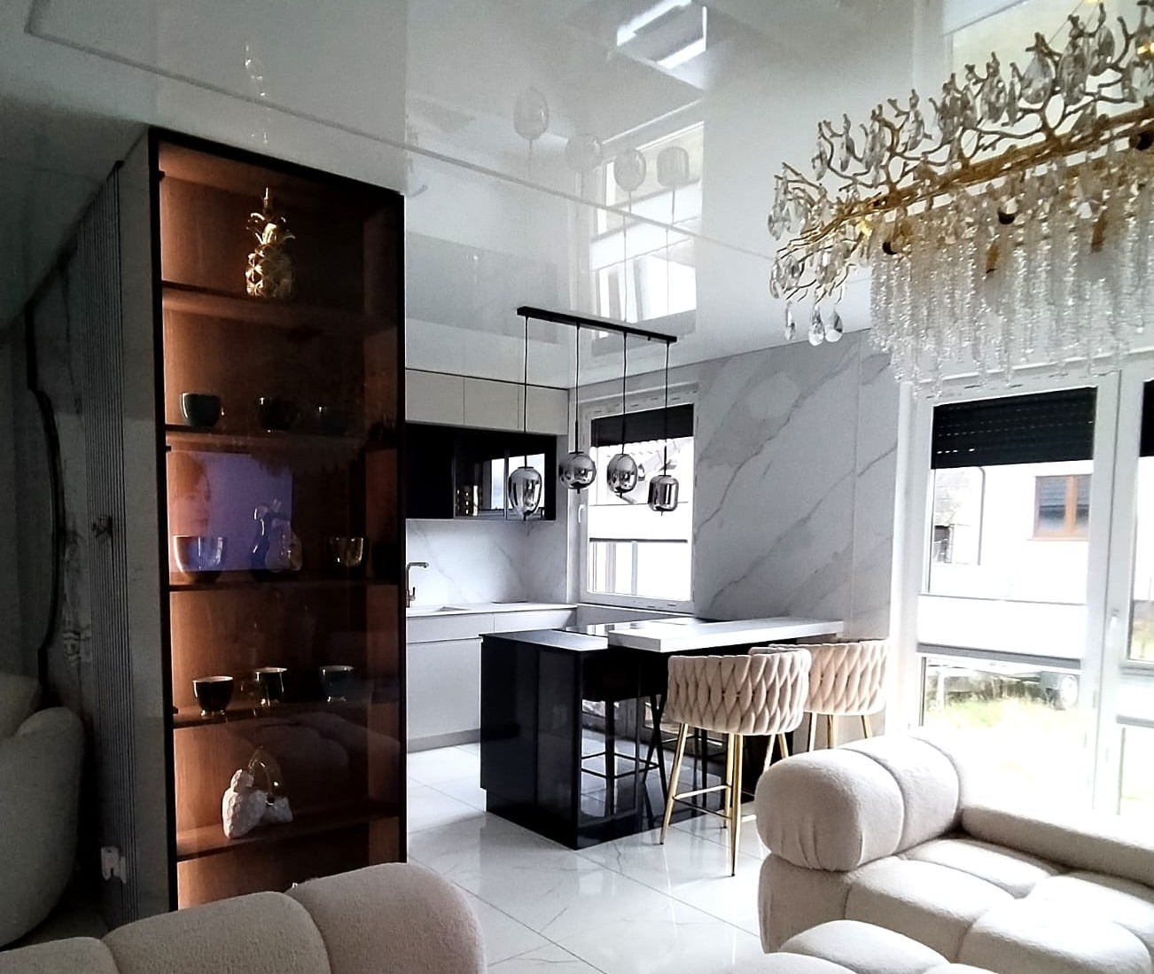 Jasna kuchnia z połyskiem: gładkie fronty bez uchwytów, podświetlenie LED, przeszklona witryna, marmurowe ściany, wyspa z hokerami i ozdobny żyrandol.