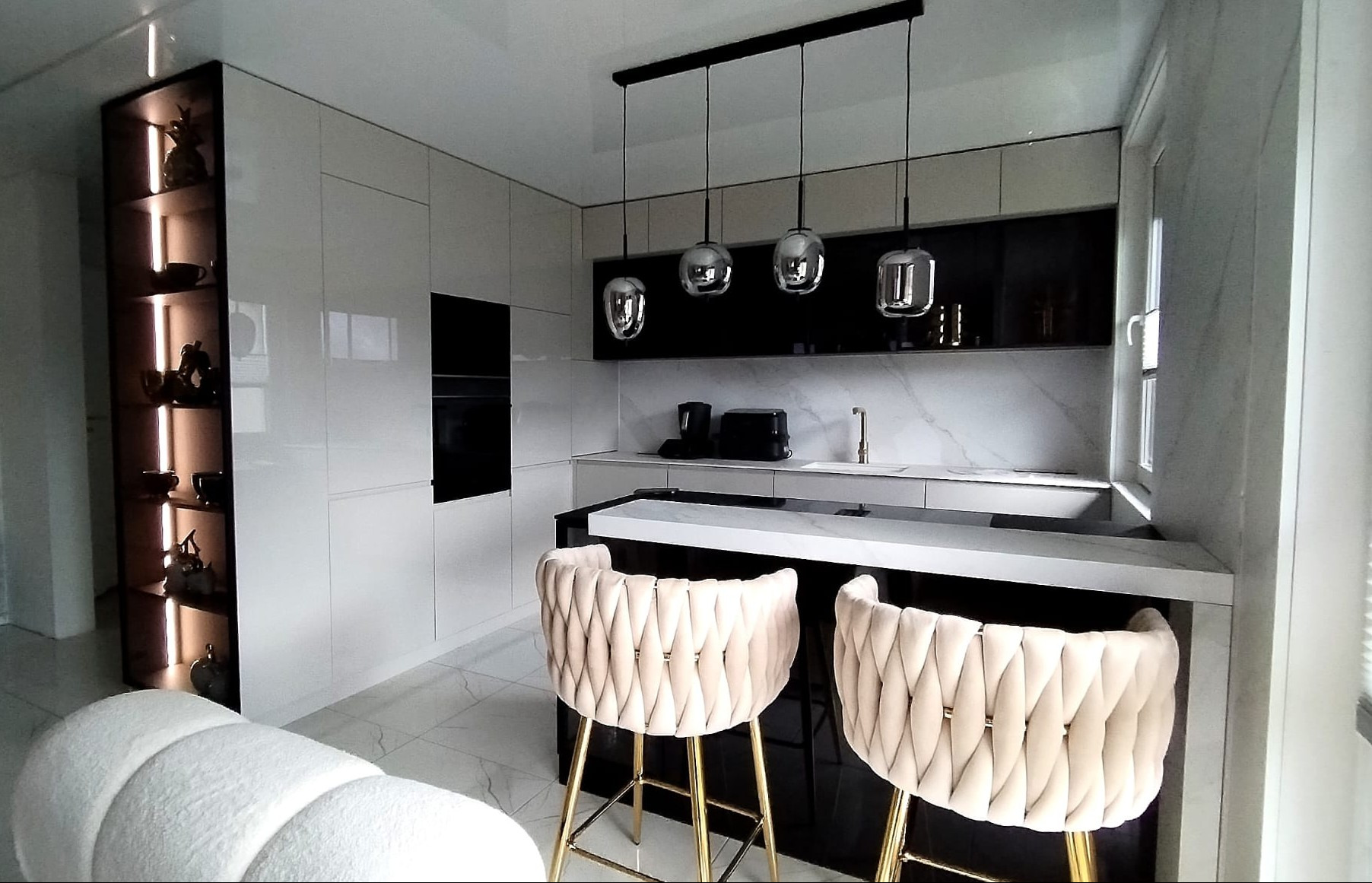 Nowoczesna kuchnia z białymi, lakierowanymi frontami, czarnymi akcentami i marmurowym blatem. Dwa beżowe hokery z pikowaniem przy wyspie. Oświetlenie punktowe i dekoracyjna półka.
