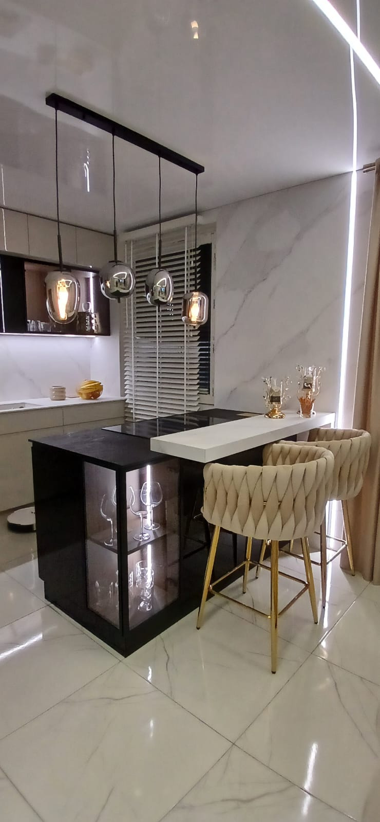 Nowoczesna kuchnia z wyspą: czarny korpus, biały blat, szklane witryny z kieliszkami, beżowe hokery na złotych nogach, marmurowe ściany i podłoga, designerskie lampy.