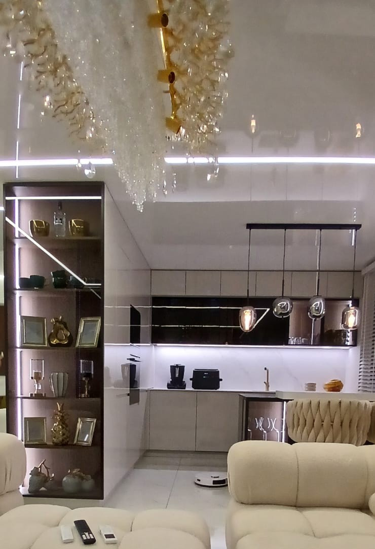 Nowoczesna kuchnia z oświetleniem LED, szklanymi półkami i dekoracyjnym żyrandolem. Widoczny robot sprzątający na podłodze i fragment sofy w kolorze kremowym.