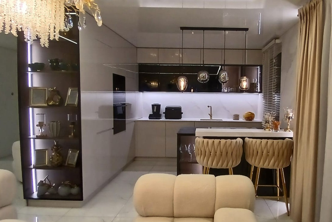 Elegancka kuchnia z wyspą i hokerami w odcieniach beżu, oświetlona wiszącymi lampami. Na półkach dekoracje, a w tle marmurowa ściana i zasłony.