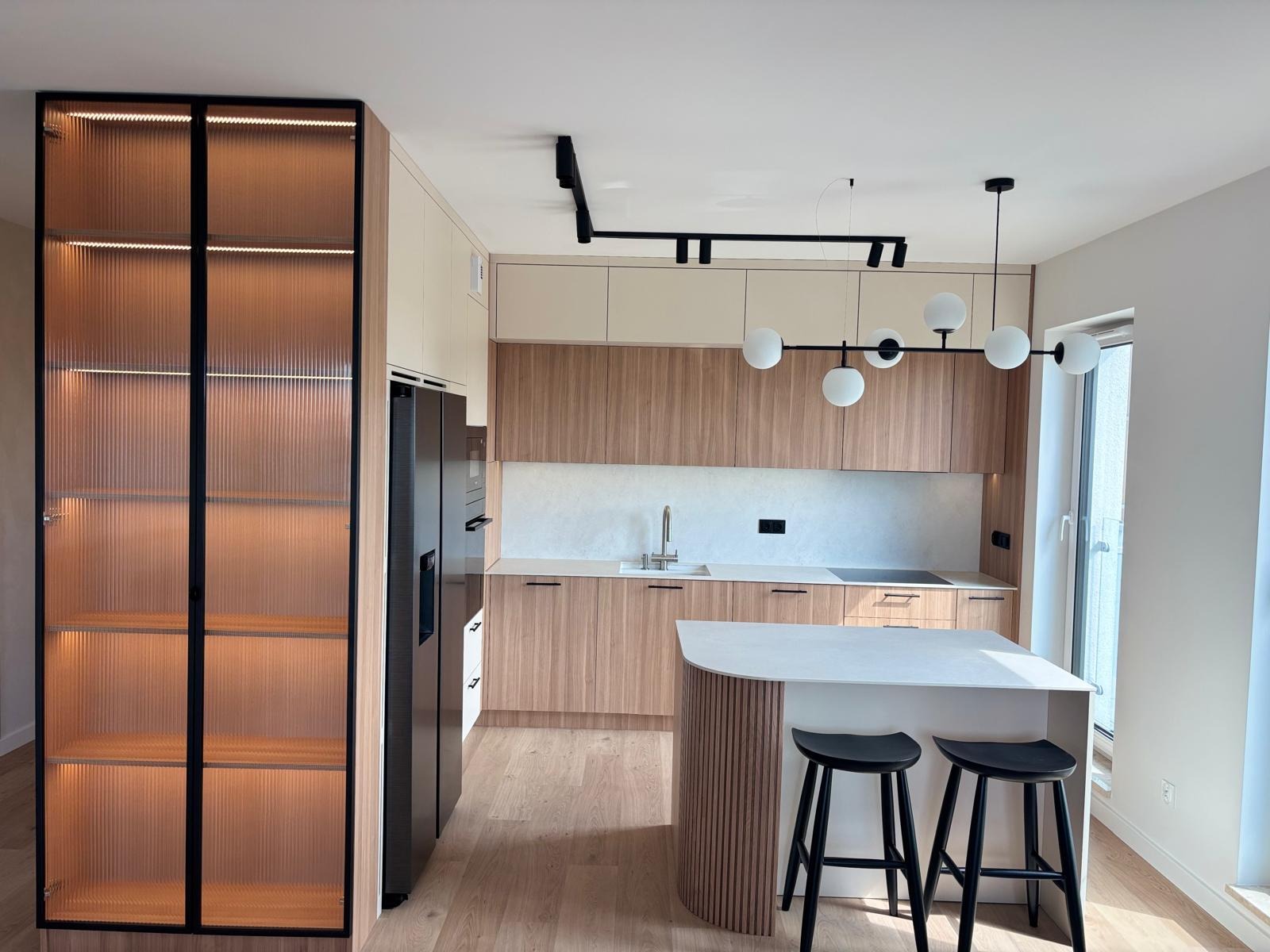 Nowoczesna kuchnia z drewnianymi frontami, wyspą i szklaną witryną z podświetleniem LED. Minimalistyczny design, czarne akcenty oświetlenia i stołków barowych.