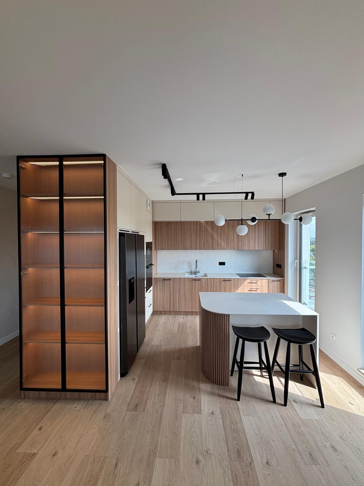 Nowoczesna kuchnia z drewnianymi frontami, wyspą z białym blatem i hokerami. Na ścianie szafki i oświetlenie. Charakterystyczna witryna z ryflowanego szkła.