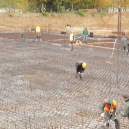 Rozległa siatka zbrojeniowa na budowie, pracownicy w kaskach i kamizelkach odblaskowych, dźwig z łańcuchami. Widok na przygotowanie fundamentów pod budynek.