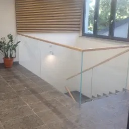 Szklana balustrada schodowa z drewnianą poręczą i subtelnym oświetleniem punktowym, widok z góry schodów w nowoczesnym wnętrzu.