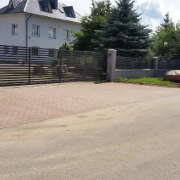 Nowoczesna, pozioma brama wjazdowa z metalowymi przęsłami i słupkami z szarej cegły, prowadząca do budynku firmy z widocznym logo, otoczona brukowaną nawierzchnią i zielenią.