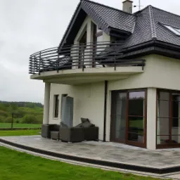 Nowoczesny dom w Kościerzynie z balkonem zabezpieczonym czarną, metalową balustradą o poziomym układzie szczebli, widok z ogrodu na taras z meblami.