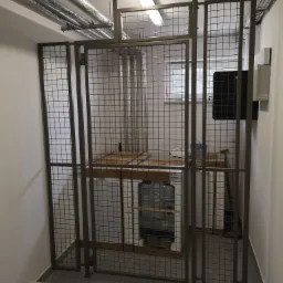 Metalowa klatka zamykająca dostęp do urządzeń technicznych w pomieszczeniu, widoczne rury wentylacyjne pod sufitem i skrzynka elektryczna na ścianie.