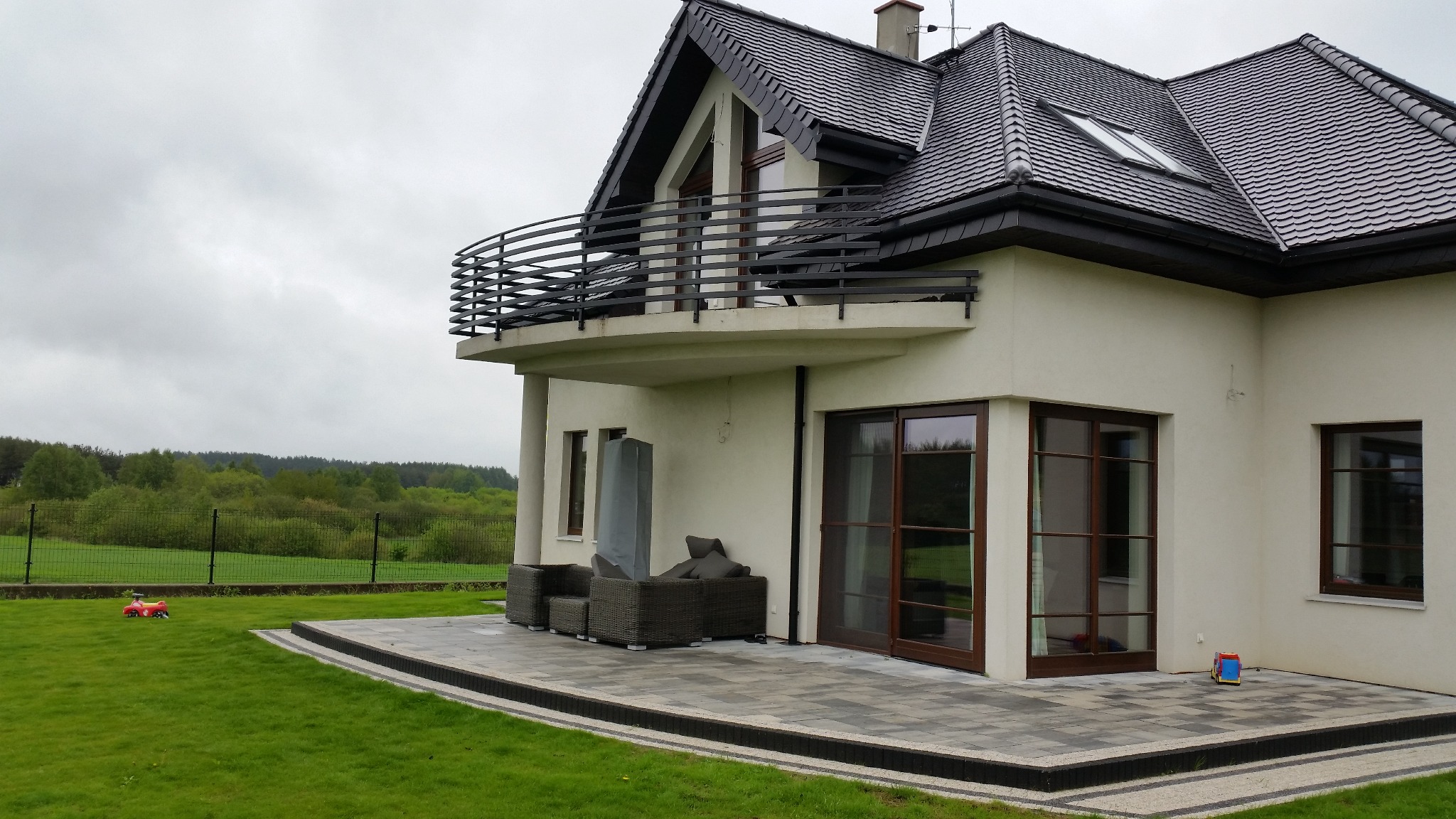 Nowoczesny dom w Kościerzynie z balkonem zabezpieczonym czarną, metalową balustradą o poziomym układzie szczebli, widok z ogrodu na taras z meblami.