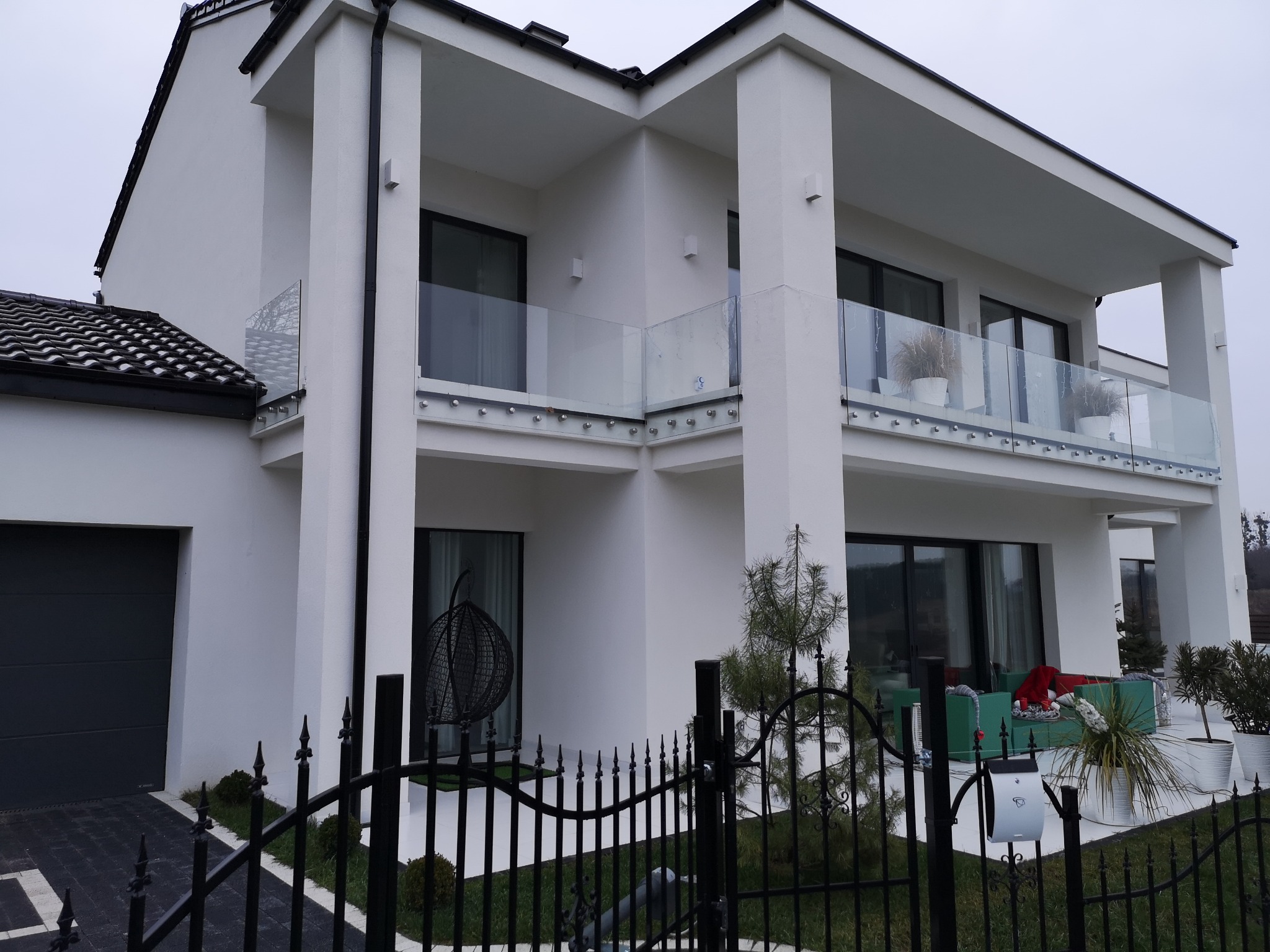 Nowoczesny, dwupiętrowy dom z białymi ścianami i szklanymi balustradami na balkonach, widoczny zza czarnego, metalowego ogrodzenia z ozdobnymi grotami.