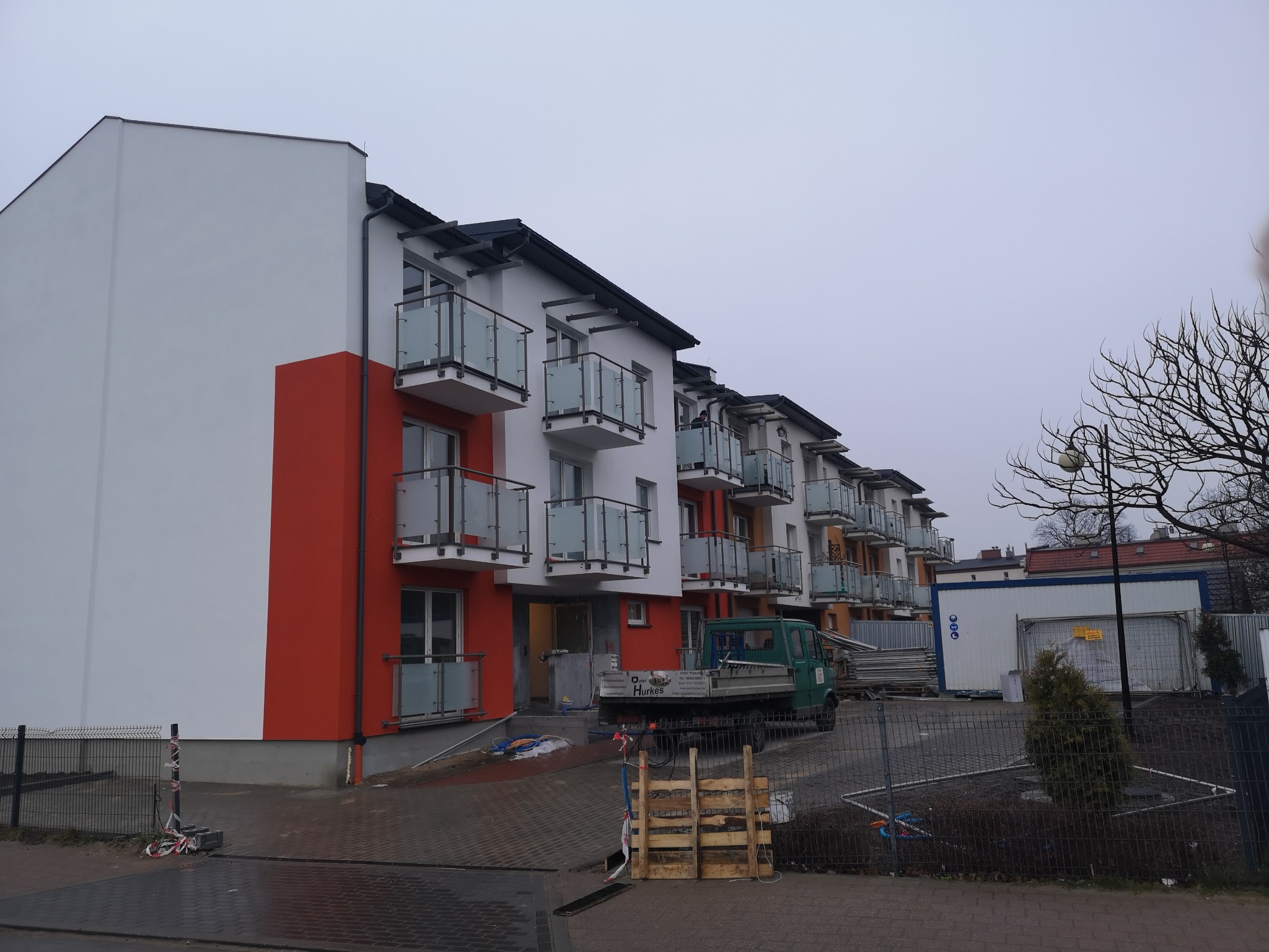 Nowe bloki mieszkalne z balkonami zabezpieczonymi szklanymi balustradami, widoczny mały samochód dostawczy i elementy placu budowy.