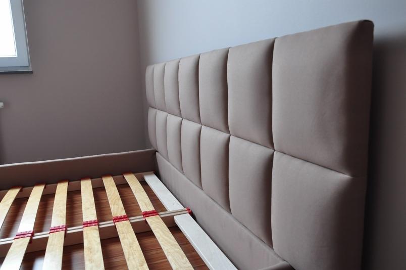 Beżowe łóżko z tapicerowanym wezgłowiem w kwadratowe panele, widoczny stelaż z drewnianych listew i czerwonymi mocowaniami, ustawione w pokoju z jasnymi ścianami.