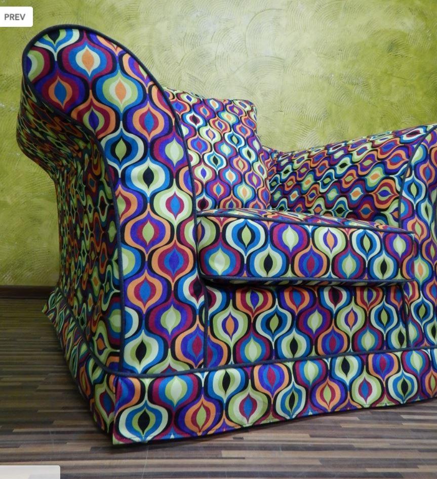 Krzesło tapicerowane w stylu retro z wzorem w kolorowe łezki, z ciemną lamówką, na tle oliwkowej ściany i drewnianej podłogi.