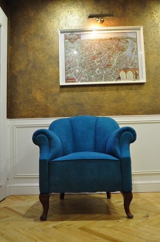 Turkusowy fotel typu uszak na drewnianych nogach w pomieszczeniu z drewnianą podłogą w jodełkę, na ścianie mapa Krakowa w białej ramie i lampka.