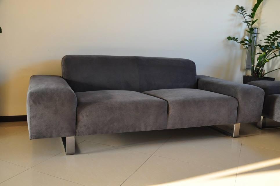 Grafitowa sofa dwuosobowa z podłokietnikami, na metalowych nogach, z rośliną doniczkową w tle.