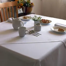 Biały, gładki obrus na stole nakrytym do śniadania, z widocznymi elementami zastawy: dzbankiem, filiżanką, mlecznikiem, sztućcami i talerzykami z pieczywem.