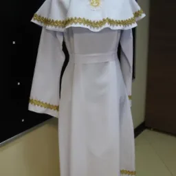 Biała alba komunijna z pelerynką, zdobiona złotą koronką i symbolem IHS, prezentowana na manekinie.