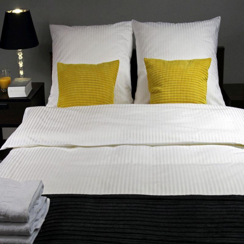 Starannie zaścielone łóżko z białą pościelą w prążki, dwoma żółtymi poduszkami z fakturą w kratkę, stosem białych ręczników i czarną narzutą, obok lampka nocna z czarnym abażurem i szklaną podstawą.