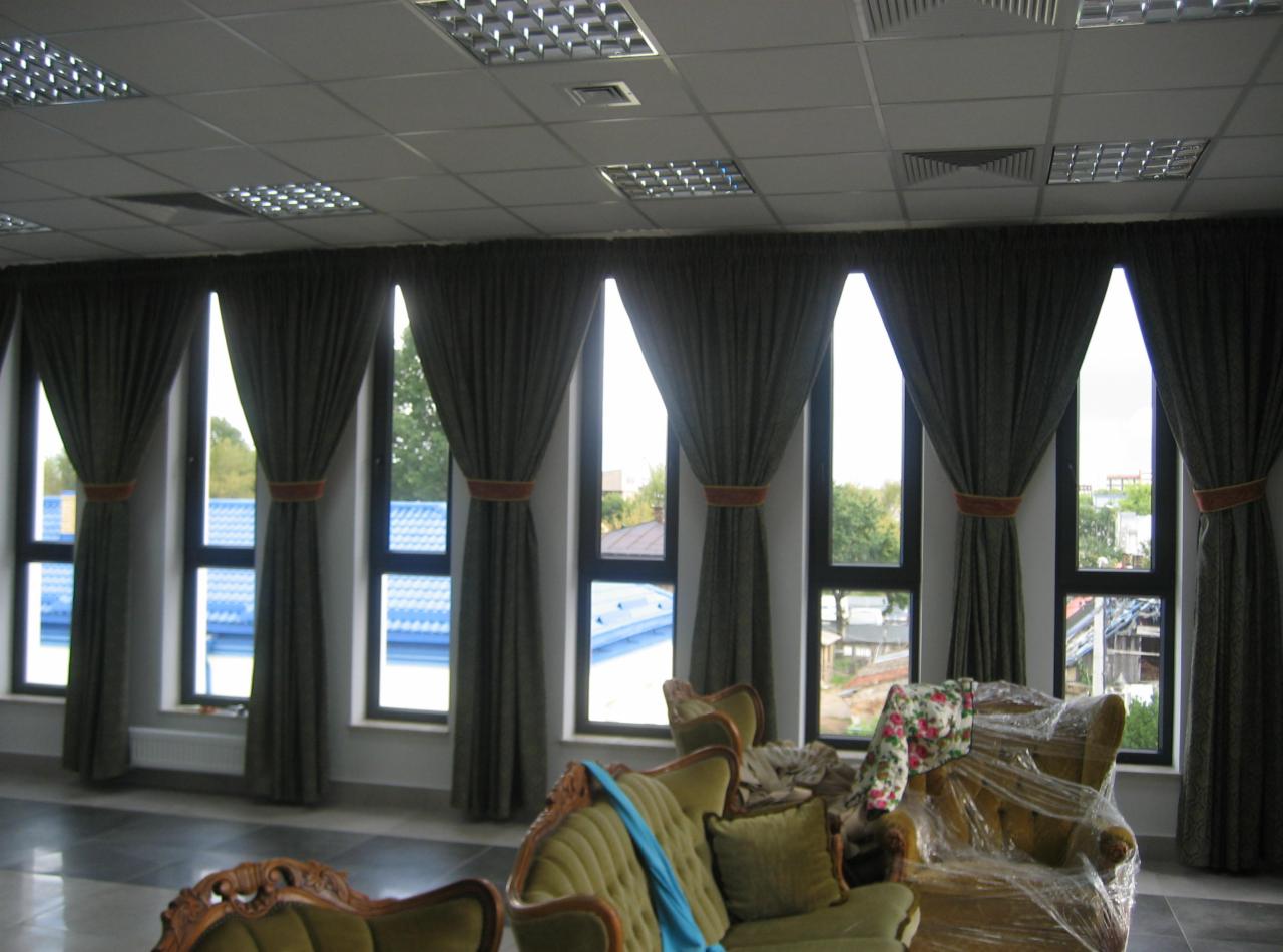 Szare, drapowane zasłony z pomarańczowymi akcentami zawieszone w wysokich oknach, widok na zewnątrz z fragmentami zabudowań, w tle widoczne krzesła w stylu retro, jedno z nich owinięte folią.