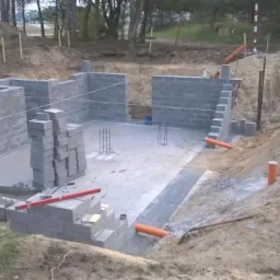 Budowa fundamentów z bloczków betonowych. Częściowo wymurowane ściany, widoczne zbrojenia, poziomica i pomarańczowe rury wentylacyjne. Wykop otoczony ziemią.