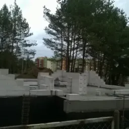 Fundamenty domu z keramzytu w trakcie budowy, widoczne białe bloczki i izolacja fundamentów, w tle drzewa iglaste i budynek mieszkalny.