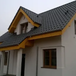 Biały dom z ciemną dachówką i drewnianymi elementami wykończeniowymi. Widoczne okna i fragment sąsiedniego budynku. Pochmurne niebo w tle.