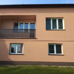 Dwupiętrowy dom z balkonem i beżową elewacją, widoczne zacieki na ścianie.