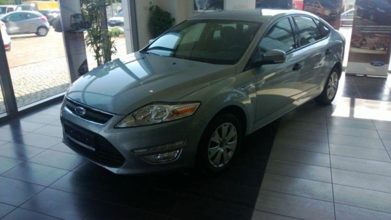 Jasnoszary Ford Mondeo sedan, prezentowany w salonie samochodowym z widoczną rośliną doniczkową przy oknie i plakatami reklamowymi w tle.