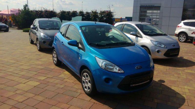 Turkusowy Ford Ka na tle salonu samochodowego, zaparkowany na kostce brukowej obok szarego i srebrnego egzemplarza.