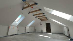 Białe poddasze ze skosami i oknami dachowymi. Widoczne drewniane elementy konstrukcyjne i świeżo wylana posadzka. Minimalistyczne wykończenie, surowy stan.