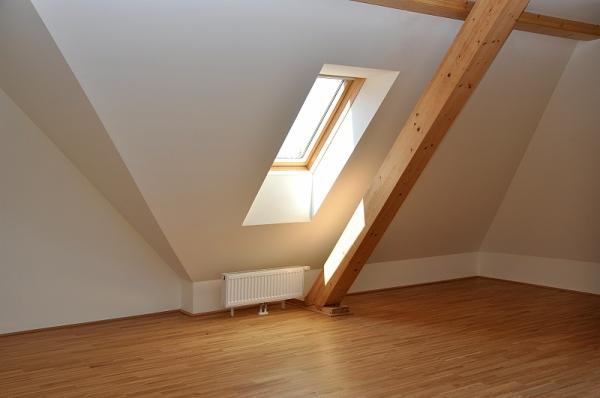 Jasne poddasze z drewnianą podłogą i belką konstrukcyjną. Okno dachowe wpuszcza światło na białą ścianę. Widoczny grzejnik.