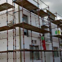 Ocieplanie elewacji domu jednorodzinnego ze styropianem, widoczne rusztowania i pracownik w czerwonych spodniach.