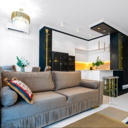 Szymon Stach - Salon z kuchnią w stylu glamour: beżowa sofa z dekoracyjną poduszką, białe meble kuchenne z czarnymi akcentami i złotymi lampami, marmurowa podłoga.