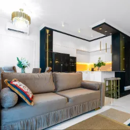 Salon z kuchnią w stylu glamour: beżowa sofa z dekoracyjną poduszką, białe meble kuchenne z czarnymi akcentami i złotymi lampami, marmurowa podłoga.