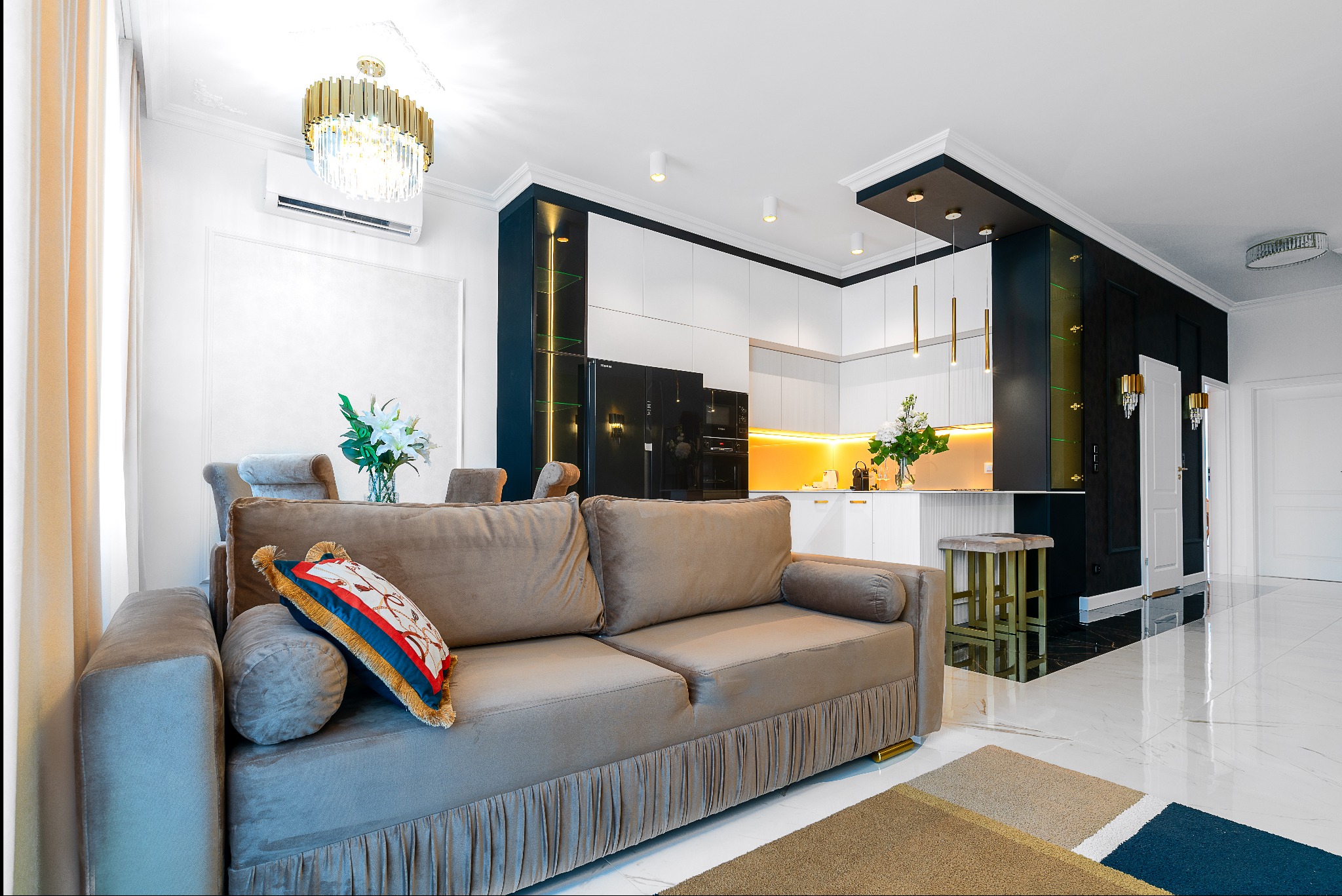 Salon z kuchnią w stylu glamour: beżowa sofa z dekoracyjną poduszką, białe meble kuchenne z czarnymi akcentami i złotymi lampami, marmurowa podłoga.