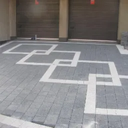 Szara kostka brukowa z białym wzorem geometrycznym przed garażem, widoczna butelka z wodą.