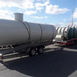 Transport szamba betonowego na przyczepie lawetą, widoczne pasy zabezpieczające ładunek i dodatkowe elementy szamba z tyłu.