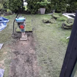 Przygotowanie podłoża pod trawnik w Szczawnie-Zdroju: ubijarka spalinowa zagęszcza ziemię w wykopie, obok leżą zwinięte rolki darni, w tle tuje i donice z roślinami.