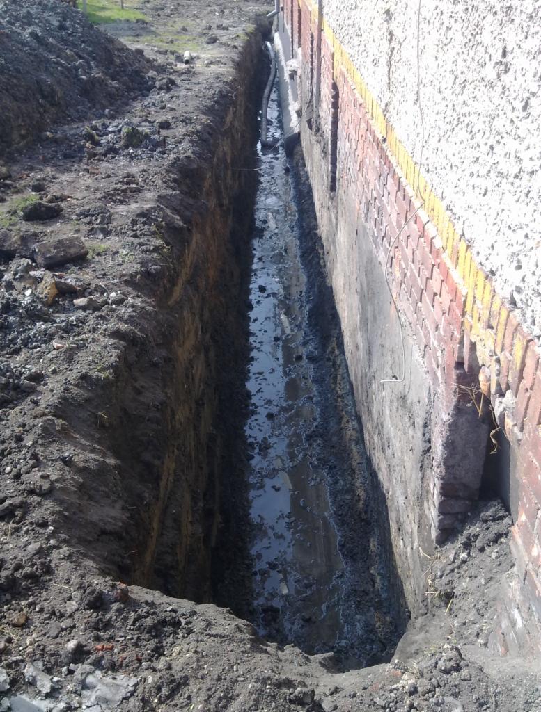 Wykop wzdłuż fundamentu budynku z widocznymi rurami. Prace ziemne przygotowawcze pod instalacje wod-kan. Grunt gliniasty, ściana budynku z cegły i betonu.