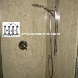 Kabina prysznicowa wykończona jasnym kamieniem naturalnym z widoczną armaturą i logo firmy kamieniarskiej. Minimalistyczny design, kamienne wykończenie ścian.