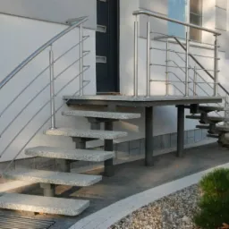 Granitowe schody zewnętrzne z metalową balustradą prowadzące do drzwi wejściowych budynku, z widocznym fragmentem elewacji i zieleni w tle.